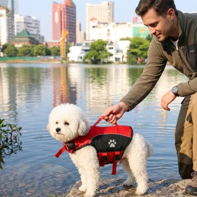 Homme tenant la poignée du gilet de sauvetage pour chien rouge porté par un Bichon frisé au bord de l'eau.