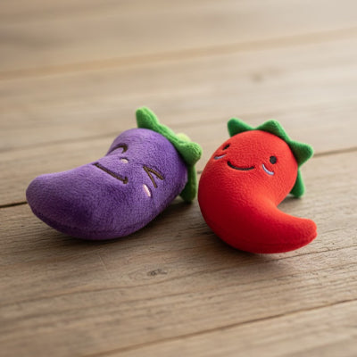 Un jouet chien aubergine et un jouet chien piment rouge en peluche posés sur du bois.