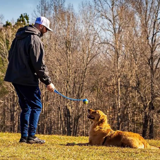 Homme à casquette avec lanceur de balle pour chien bleu face à un Golden Retriever couché.