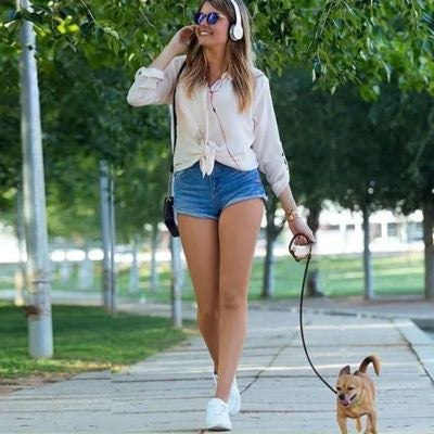 Jeune femme avec casque audio promenant un petit chien roux avec une longe chien fine en cuir.
