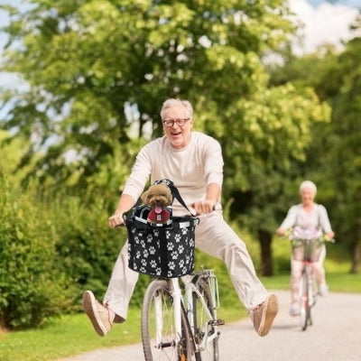 Homme âgé à vélo avec un caniche marron dans un panier chien vélo noir à motifs pattes dans un parc.