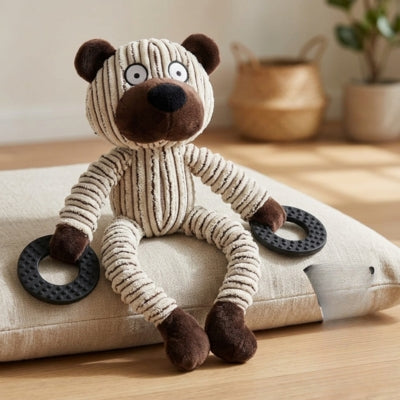 Peluche pour chien en forme d'ours rayé avec anneaux noirs posée sur un grand coussin beige.