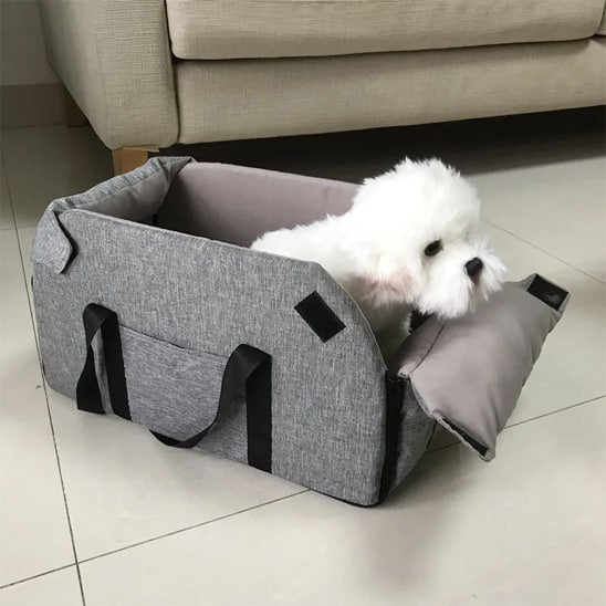 Bichon blanc dans un siege auto chiens gris dézippé servant de panier au sol près d'un canapé.
