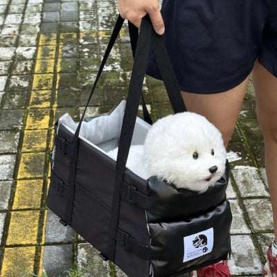 Un bichon blanc transporté à la main dans son siege auto pour chiens noir transformé en sac.